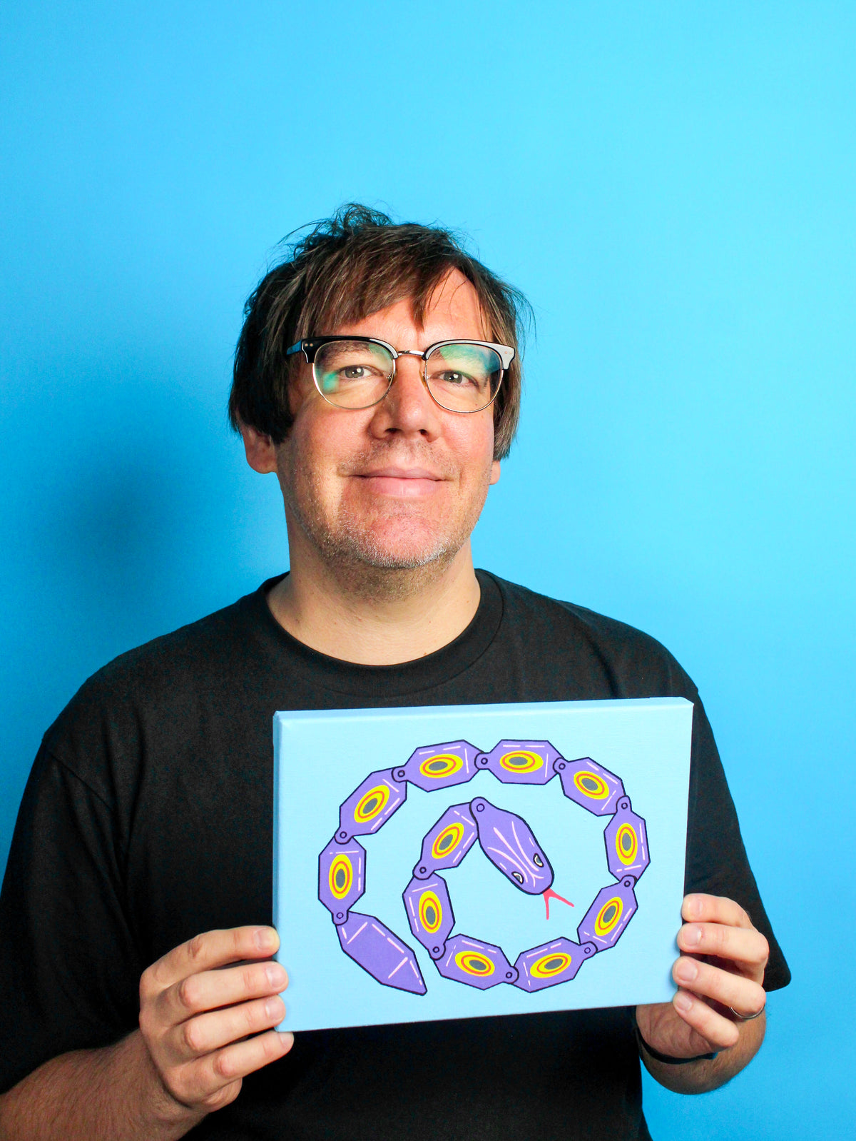A man with glasses and a black t-shirt holds up a square canvas painting depicting a purple and yellow snake-like chain pattern on a light blue background, set against a solid blue backdrop.